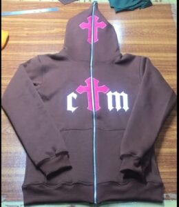 "Flat-lay of a brown zip-up hoodie with a bold pink cross on the hood and center chest, flanked by white gothic-style 'ctm' lettering. Displayed on a wooden surface, the hoodie features ribbed cuffs, dual front pockets, and a full-length zipper."