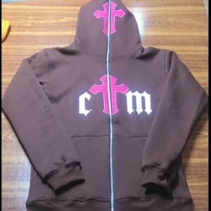 "Flat-lay of a brown zip-up hoodie with a bold pink cross on the hood and center chest, flanked by white gothic-style 'ctm' lettering. Displayed on a wooden surface, the hoodie features ribbed cuffs, dual front pockets, and a full-length zipper."
